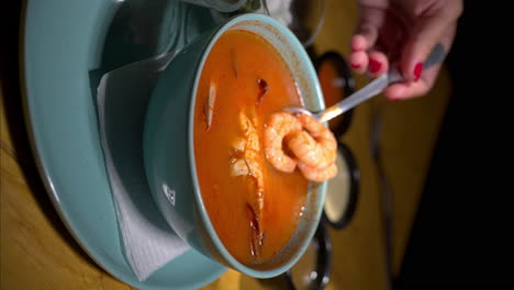 Vertical-slow-motion-of-a-female-hand-holding-shrimp-with-a-spoon-out-of-a-seafood-red-soup-with-a-crab-on-a-blue-bowl
