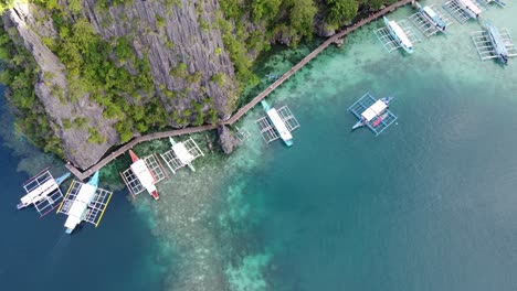 Tour-boats-in-crystal-blue-water-and-jetty-boardwalk-along-sheer-karst-cliffs-of-Kayangan-lake,-Coron-island