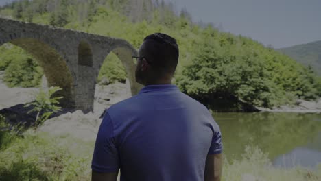 Panning-around-a-tourist-who-is-admiring-the-architectural-design-of-the-Devil's-Bridge-and-beautiful-scenery-of-Arda-River-right-next-to-the-Rhodope-Mountains-in-Bulgaria