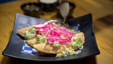 Slow-motion-close-up-of-traditional-mexican-empanadas-with-chopped-lettuce-and-red-onion-on-top-and-someone-pouring-sour-cream-on-them