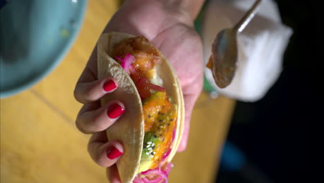 Vertical-slow-motion-of-a-female-woman-holding-a-mexican-traditional-taco-with-avocado-and-red-onion,-pouring-red-hot-salsa-on-it