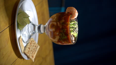 Vertikale-Aufnahme-Eines-Traditionellen,-Typisch-Mexikanischen-Meeresfrüchte-Cocktails-Mit-Garnelen-Und-Avocado,-Serviert-Auf-Einer-Glasschale-Mit-Crackern-Und-Limettenscheiben