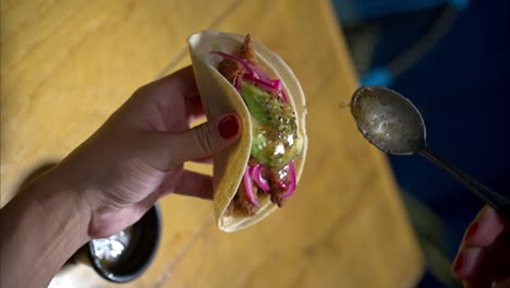 Vertical-slow-motion-of-a-woman-holding-a-shrimp-seafood-taco-with-red-onion-and-avocado-serving-pouring-green-salsa-sauce-on-top-at-a-traditional-mexican-restaurant