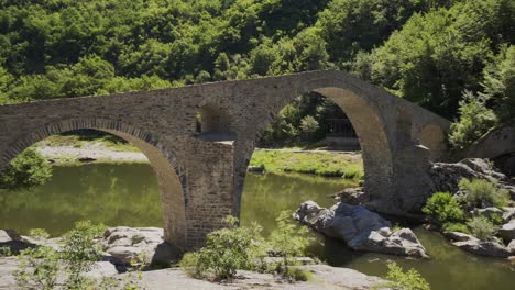 Panning-from-the-left-to-the-right-side-of-the-Devils-bridge-and-Arda-River,-situated-at-the-foot-of-Rhodope-Mountains-in-Ardino,-Bulgaria