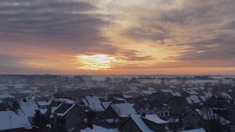 Aerial-pullback-above-snow-covered-suburban-roofs-with-fire-yellow-sunset-streak-in-sky-above-misty-fog
