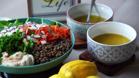 Linsensalat-Auf-Rotierender-Plattform,-Um-Einen-Salat-Zuzubereiten,-Hummus-Mit-Spinat-Und-Karottenscheiben-Auf-Den-Teller-Zu-Geben,-Korianderzwiebeln,-Mandelscheiben,-Dressing-Und-Zitronen