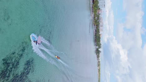 Vertical-drone-shot-of-tourist-on-banana-boat-pulling-by-motorboat-on-Caribbean-sea