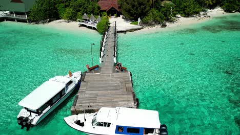 Tropical-Island-Paradise-found---Malahini-Kuda-Bandos,-Maldives:-Aerial-dronie-from-jetty---guests-waving,-island-reveal