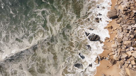 Aerial-top-down-shot-of-crashing-waves-arriving-sand-and-rocks-of-Nazare-Beach-in-summer---circling-shot