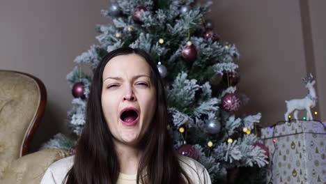 Tired-woman-yawns-near-decorated-artificial-Christmas-tree-and-gift-box