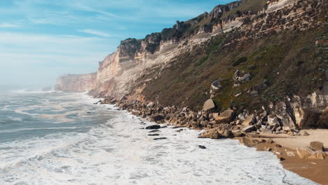 Aerial-backwards-shot-of-foamy-waves-reaching-sandy-beach-of-Nazare-with-gigantic-cliff-Formation-in-background
