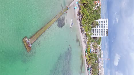 Vertical-drone-shot-of-Tourist-walking-on-jetty-at-Playa-Boca-Chica-at-sandy-beach-with-palm-trees-in-summer---Santo-Domingo,-Dominican-Republic