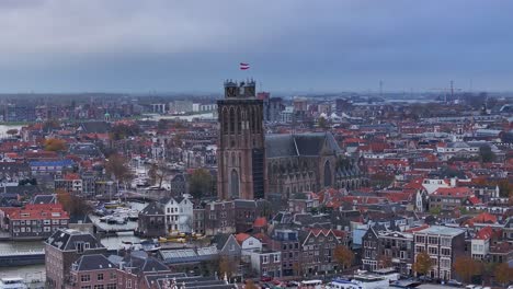 Flag-of-Netherlands-flying,-Church-shrine-above-the-rooftops-of-Dordrecht