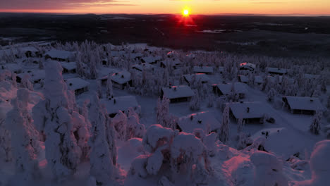 Aerial-view-around-snowy-trees,-revealing-mountain-cottages,-winter-sunset-in-Lapland