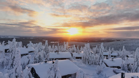Aerial-view-low-over-a-snowy-cabins-toward-arctic-wilderness,-sunrise-in-Lapland