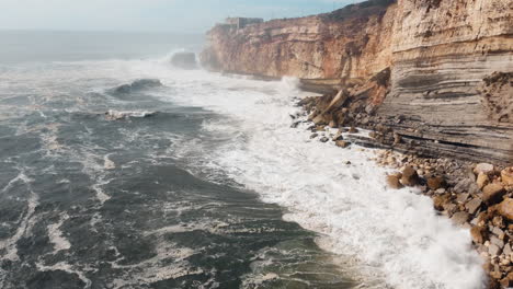 Aerial-of-rocky-beach-with-crashing-waves-in-Nazare,-Portugal