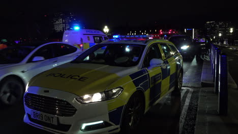 Nachts-Steht-Ein-Stadtpolizeiauto-Auf-Der-Westminster-Bridge-Geparkt,-Während-Ein-Polizeiwagen-Vorbeifährt-Und-Beamte-Im-Hintergrund-Ein-Auto-Durchsuchen