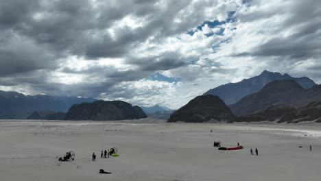 Aerial-drone-forward-moving-shot-over-paragliders-preparing-to-fly-along-Sarfaranga-desert,-surrounded-by-mountains-in-Skardu,-Pakistan-on-a-cloudy-day