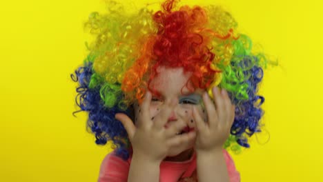 Little-child-girl-clown-in-colorful-wig-hides-behind-her-hands-and-shows-funny-faces.-Halloween
