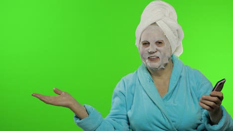 Großmutter-Im-Bademantel,-Gesichtsmaske.-Frau-Mit-Smartphone-Zeigt-Auf-Etwas