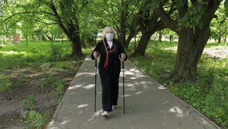 Active-senior-old-woman-in-mask-training-Nordic-walking-in-park-at-quarantine