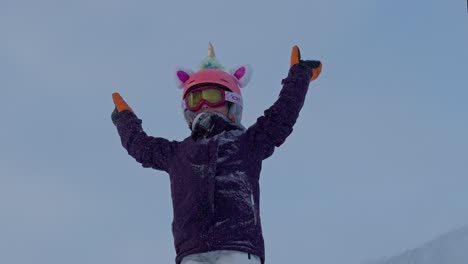 7-year-old-girl-with-unicorn-helmet-raise-hands,-cheering-on-top-of-hill-while-snowing,-slow-motion