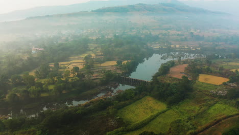 Luftaufnahme-Eines-Winterdorfes-Mit-Abgeernteten-Reisfeldern-Und-Einem-Wunderschönen-Fluss,-Der-Morgens-Mit-Nebel-In-Maharashtra-Fließt