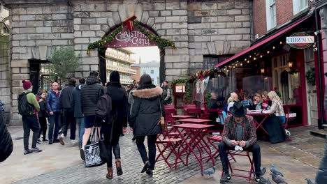 In-4K-Sitzt-Ein-Mann-Auf-Einem-Stuhl,-Während-Tauben-Auf-Seinem-Schoß-Sitzen-Und-Fressen,-Während-Passanten-Zum-Dublin-Castle-Weihnachtsmarkt-Eilen-Und-Ihn-Völlig-Ignorieren