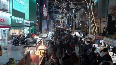 Menschenmenge,-Die-Im-Winter-Straßenessen-Auf-Dem-Myeongdong-Nachtmarkt-Isst,-Verkaufsstände-–-Hoher-Winkel