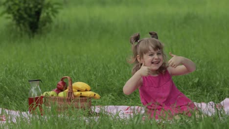 Weekend-at-picnic.-Lovely-caucasian-child-girl-on-green-grass-meadow-sit-on-blanket-show-thumbs-up