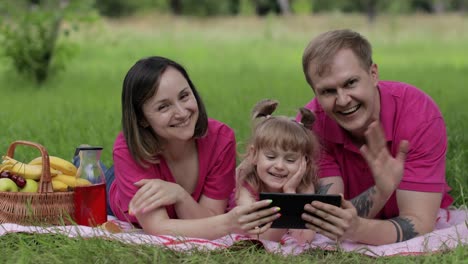 Family-weekend-picnic.-Daughter-child-girl-with-mother-and-father-play-games-on-tablet,-waving-hands