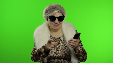 Caucasian-grandmother-woman-using-smartphone,-pointing-at-camera-with-hand