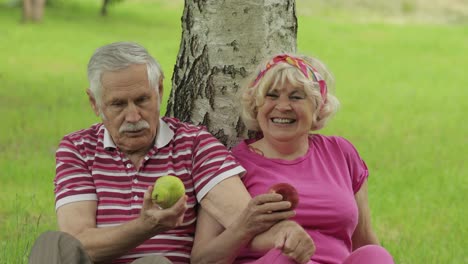 Family-weekend-picnic-in-park.-Active-senior-old-caucasian-couple-sit-near-tree-and-eating-fruits