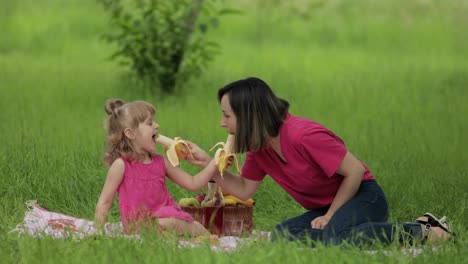 Family-weekend-at-picnic.-Daughter-child-girl-with-mother-on-grass-meadow-eating-bananas,-having-fun