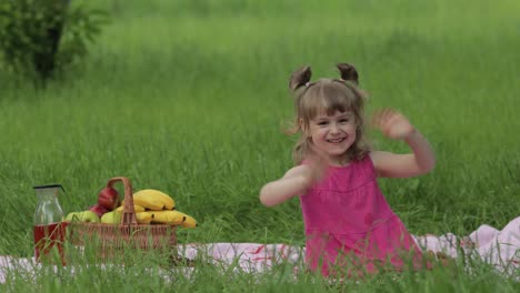 Weekend-at-picnic.-Lovely-caucasian-child-girl-on-green-grass-meadow-sit-on-blanket-waving-her-hands