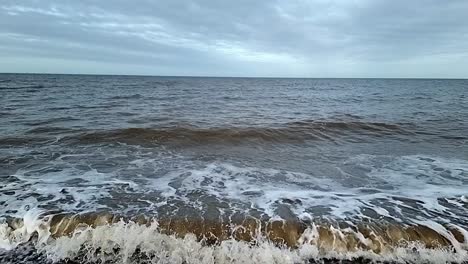 Sandy-dirty-slow-motion-tide-waves-crashing-onto-stormy-pebble-waterfront-looking-out-across-horizon