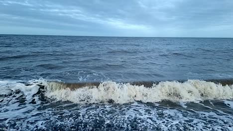 Dirty-sandy-slow-motion-tide-waves-crashing-onto-stormy-pebble-waterfront-looking-out-across-horizon