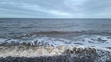 Dirty-slow-motion-tide-waves-crashing-onto-stormy-pebble-waterfront-looking-out-across-horizon