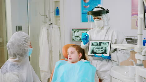 Dentist-with-face-shield-explaining-panoramic-mouth-x-ray-image-to-mother-of-kid