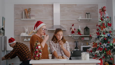 Großmutter-Schaut-Sich-Mit-Enkelin-Auf-Dem-Tablet-Ein-Online-Weihnachtsvideo-An,-Während-Großvater-Milch-Bringt