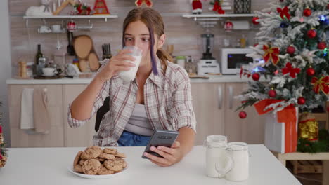 Glückliches-Mädchen-Genießt-Den-Winterurlaub,-Sitzt-Am-Tisch-In-Weihnachtlich-Dekorierter-Küche-Und-Surft-Auf-Dem-Smartphone