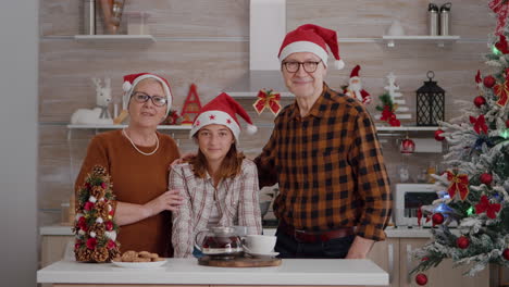 Portrait-of-happy-family-wearing-santa-hat-looking-into-camera-standing-at-table-in-xmas-decorated-kitchen