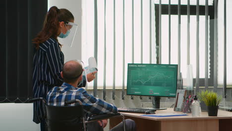 Woman-with-mask-pointing-on-desktop-talking-with-disabled-colleague
