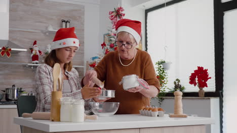 Granddaughter-using-kitchen-strainer-while-grandmother-putting-flour-ingredient