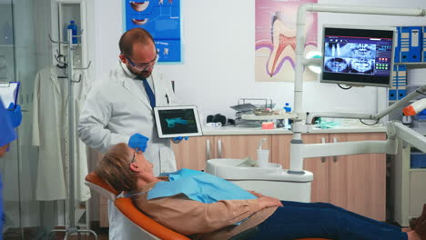 Mujer-Sentada-En-El-Sillón-Dental-Escuchando-Al-Médico-Mirando-La-Tableta