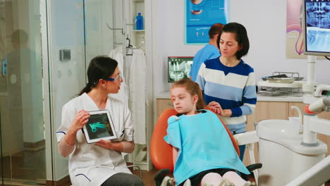 Kinder-Sitzen-Auf-Einem-Zahnarztstuhl-Und-Hören-Dem-Kinderarzt-Zu,-Der-Auf-Ein-Tablet-Schaut