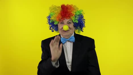 Senior-old-woman-clown-in-wig-waves-her-hands,-smiling.-Yellow-background