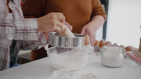 Grandmother-teaching-granddaughter-how-to-sift-flour-using-kitchen-strainer
