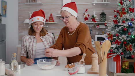 Granddaughter-with-granddaughter-breaking-egg-in-kitchen-bowl-with-flour-ingredient