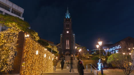 Nachtzeitraffer-Der-Myeongdong-Kathedrale-–-Menschen,-Die-In-Einem-Park,-Der-Für-Die-Weihnachtszeit-Mit-Leuchtenden-Girlanden-Geschmückt-Ist,-Stufen-Hinauf--Und-Hinuntergehen-–-Heranzoomen